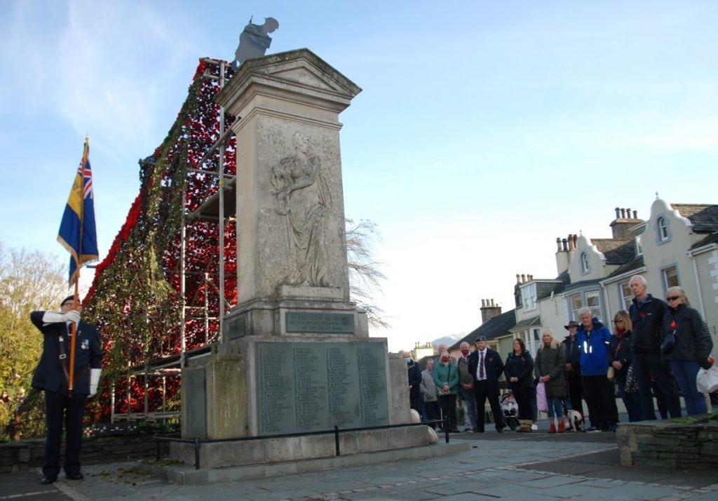 The Cenotaph in Keswick showing a cascade of poppies and crowds gathered with the British Legion standard bearer holding the flag of the local branch