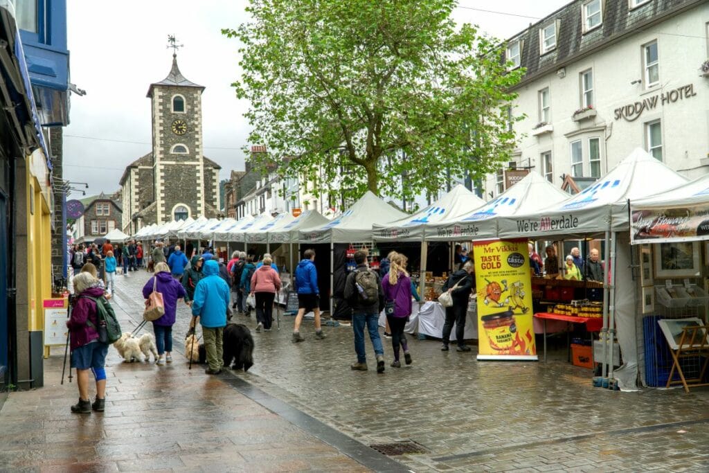 Keswick Market with the stalls on the right and shops on the left. The clock tower is in the background. The maket is very busy and the ground is wet.