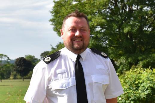 Temporary Assistant Chief Constable Jonny Blackwell stands in his white police shirt and black tie and a smile on his face. He is pictured with trees in the background 